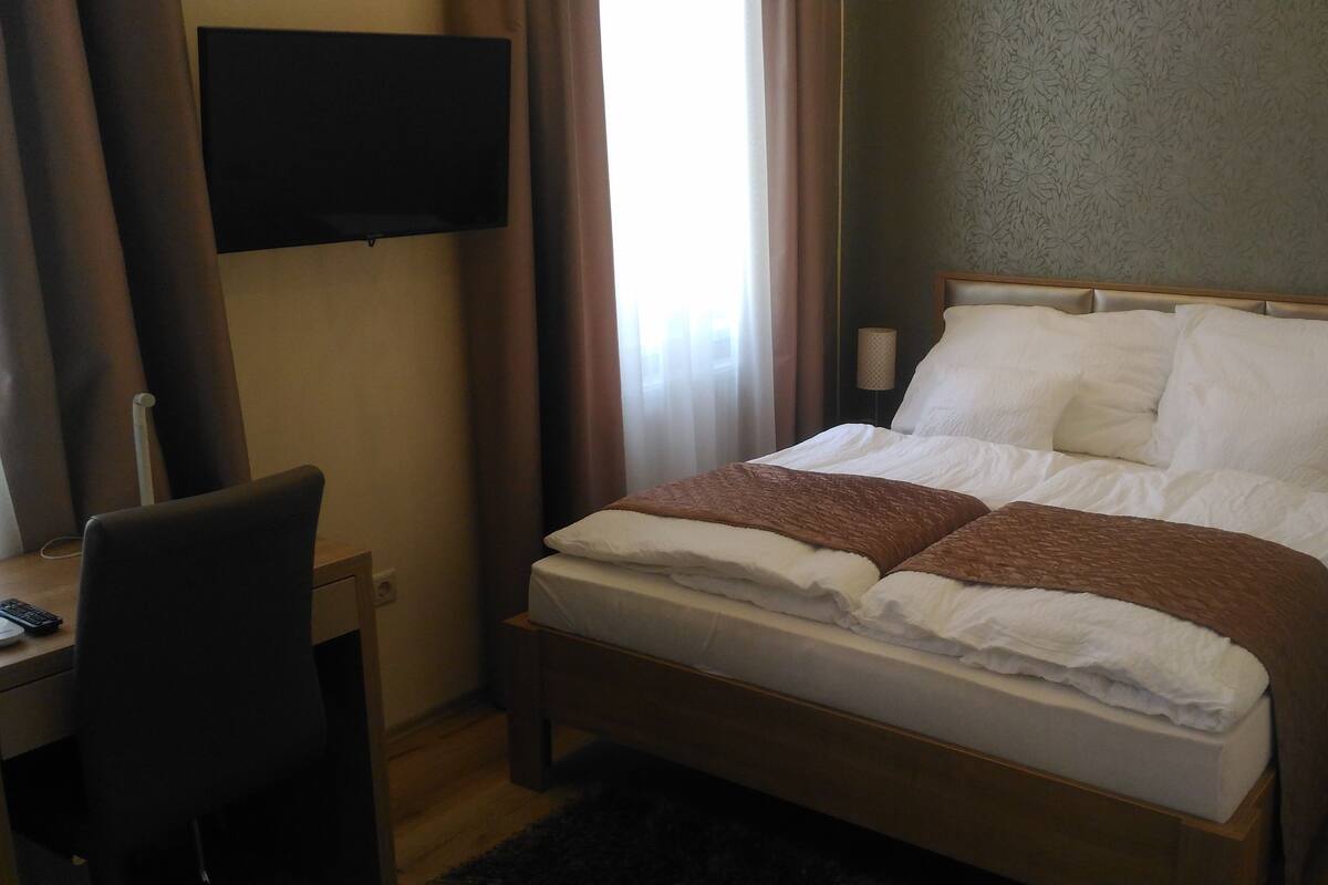 A hotel room with a bed, a desk, a chair, a TV, and a window with curtains.