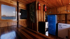 A cabin interior with wooden walls, a window with a view, a black refrigerator, a blue refrigerator, and two fire extinguishers. - Plataran Phinisi Waecicu (Labuan Bajo)