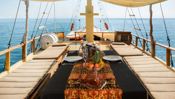 A dining table set on a boat with a view of the sea, equipped with cushions, a fruit bowl, and a decorative centerpiece. - Plataran Phinisi Waecicu (Labuan Bajo)