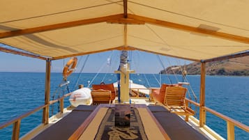 Cubierta de barco con toldo, sillas de madera, una mesa y vista al mar.