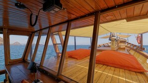 A wooden deck with a steering wheel and control panel, large windows, and orange cushions. - Plataran Phinisi Waecicu (Labuan Bajo)