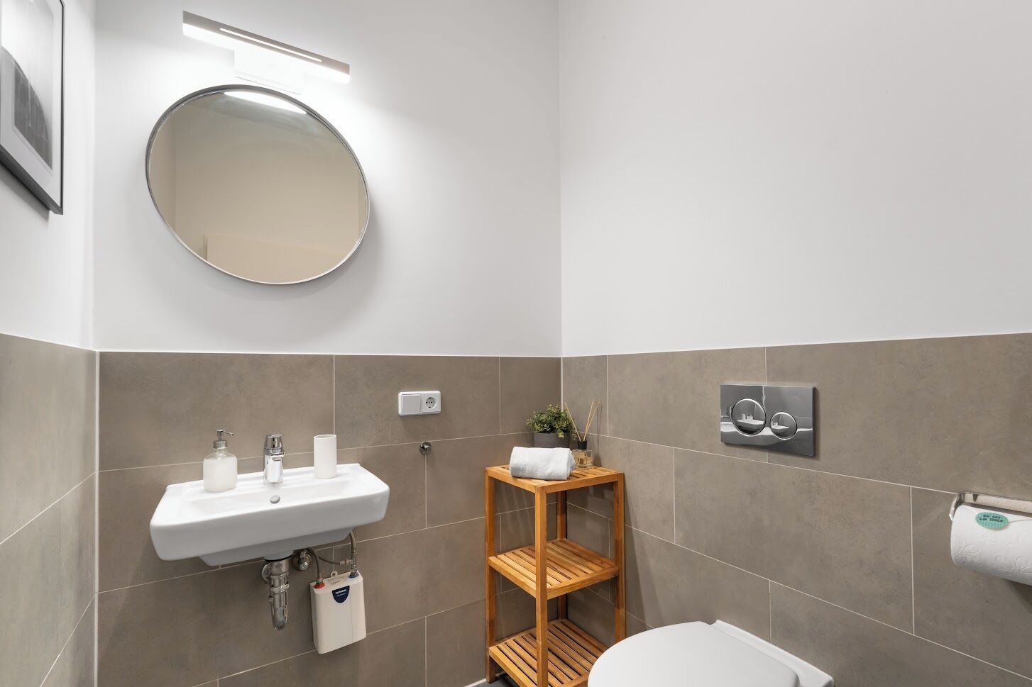 A modern bathroom with a round mirror, a sink with two faucets, a toilet, and a shower control panel.