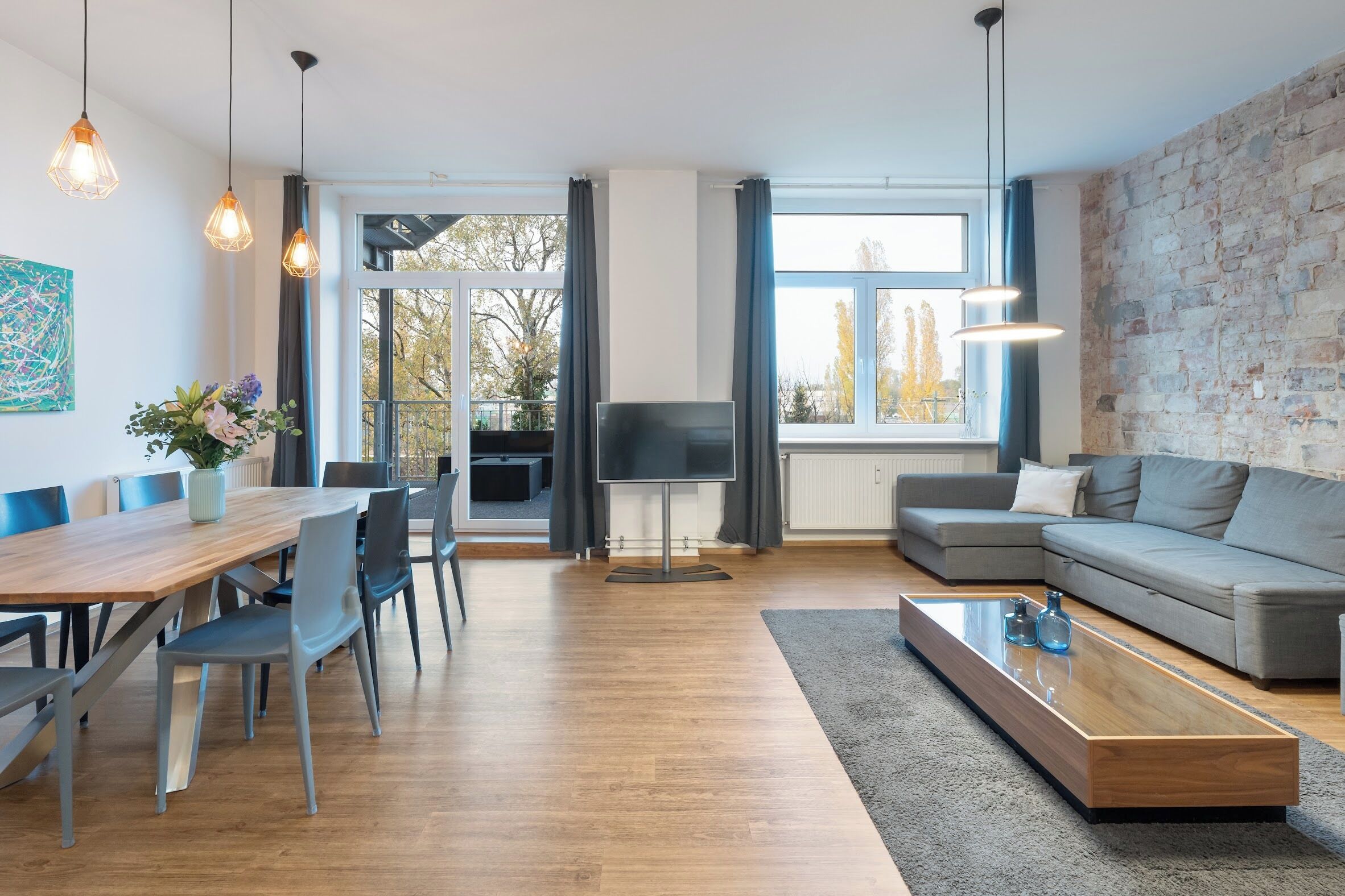 Ein modernes Wohnzimmer mit einem Holzesstisch, grauen Sofas und einem großen Fenster mit Blick auf Bäume.