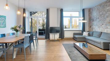 Ein modernes Wohnzimmer mit einem Holzesstisch, grauen Sofas und einem großen Fenster mit Blick auf Bäume.