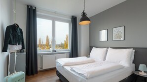 Una habitación de hotel con una cama grande, una maleta, un abrigo colgado y una ventana con vistas a árboles.