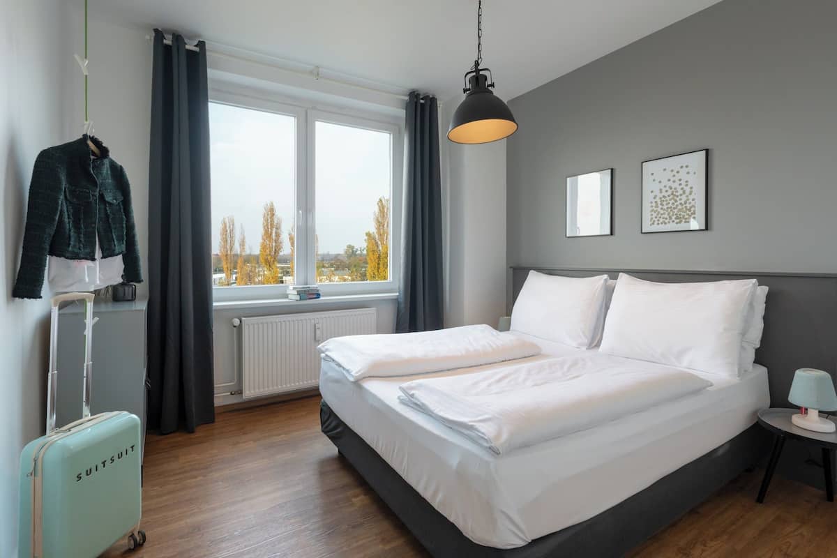 A hotel room with a large bed, a suitcase, a hanging coat, and a window with a view of trees.