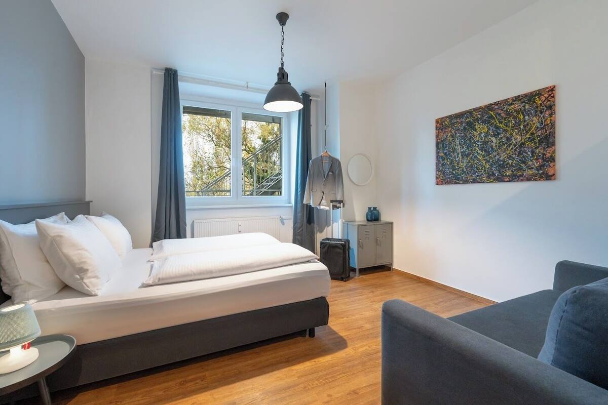 A modern bedroom with a bed, a desk, a chair, a radiator, a window with curtains, and a painting on the wall.