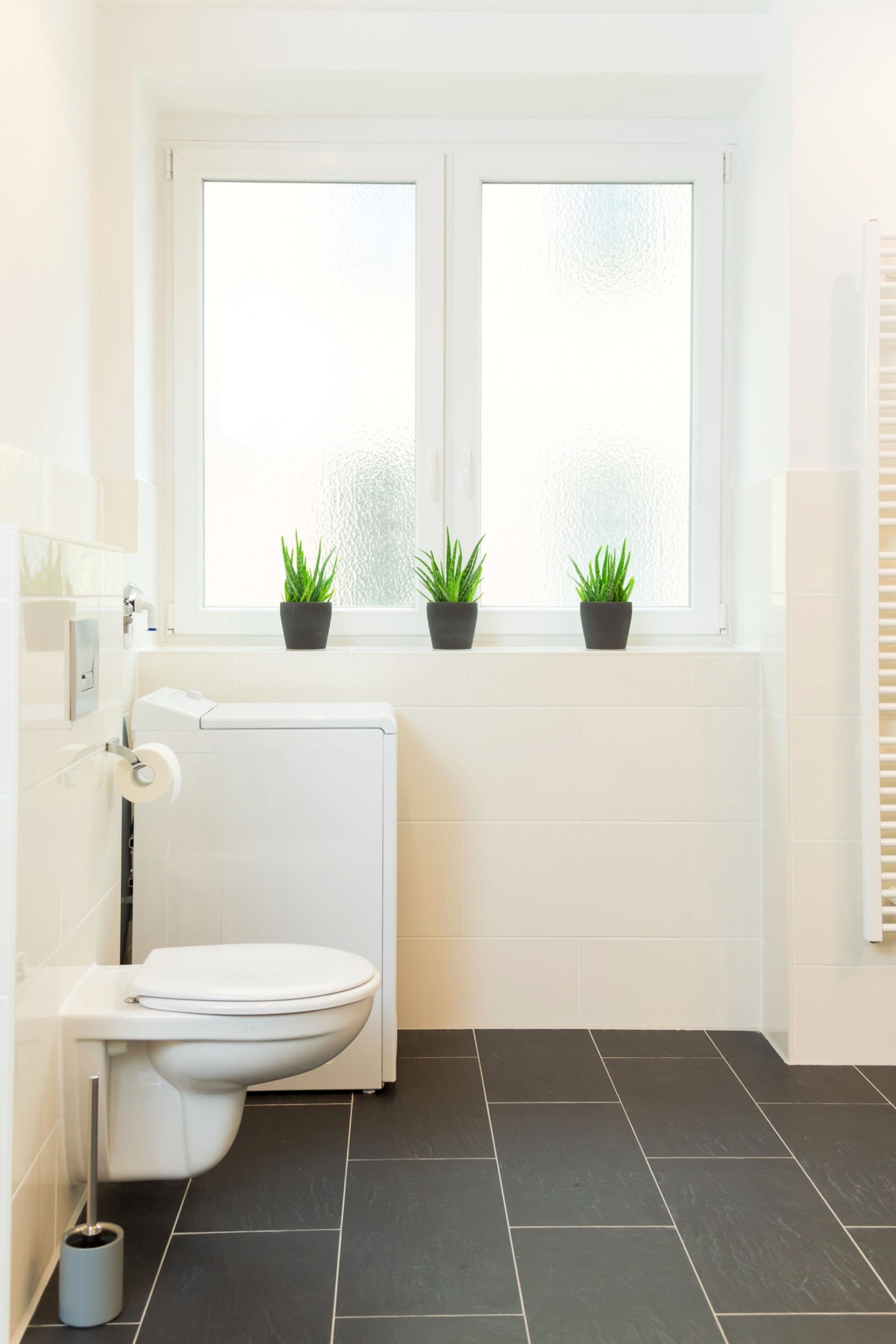 A modern bathroom with a toilet, a white radiator, and a window with three potted plants.