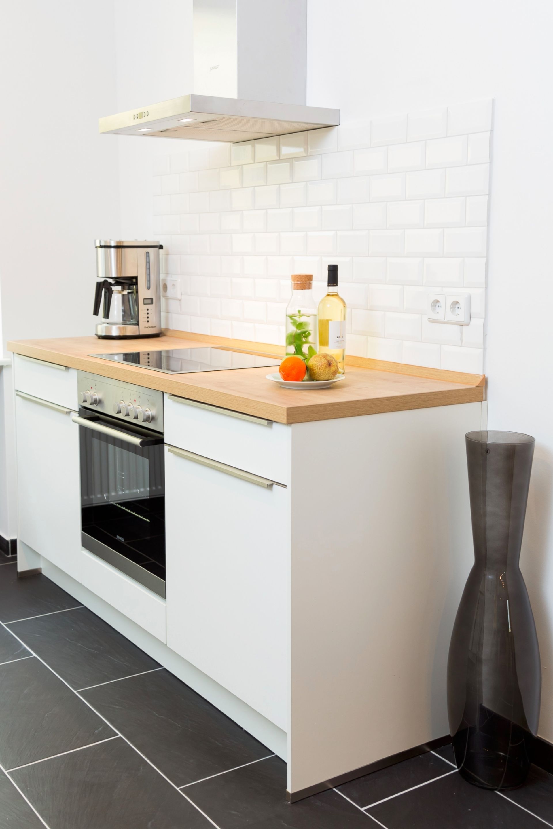 A modern kitchen with white cabinetry, a wooden countertop, an oven, and a tall vase.
