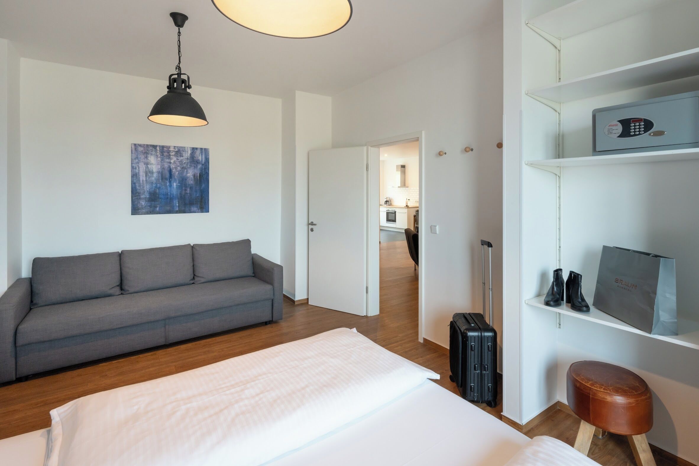 Habitación de hotel con una cama, un sofá gris, una pared blanca con un cuadro abstracto azul, una maleta negra, un taburete marrón y una estantería blanca con una caja fuerte y un bolso.