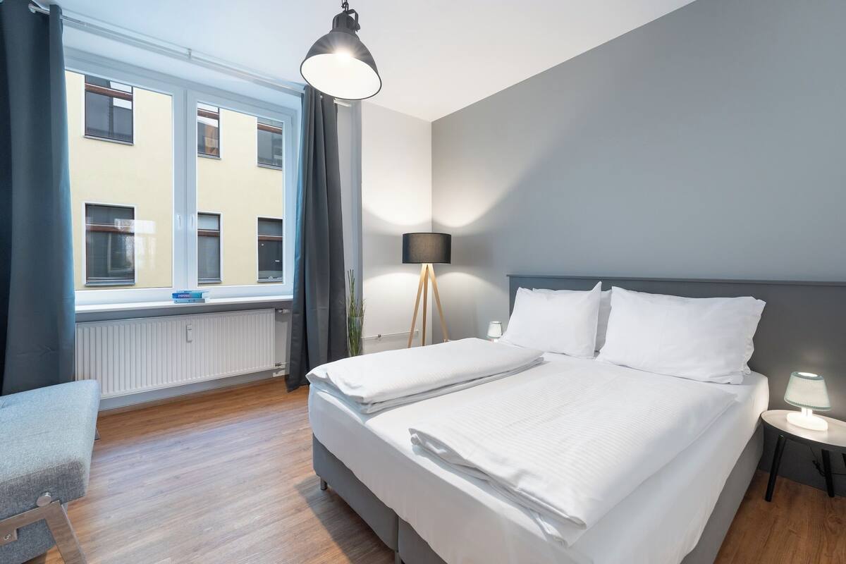 A hotel room with a large bed, bedside tables, a floor lamp, and a view of buildings through the window.