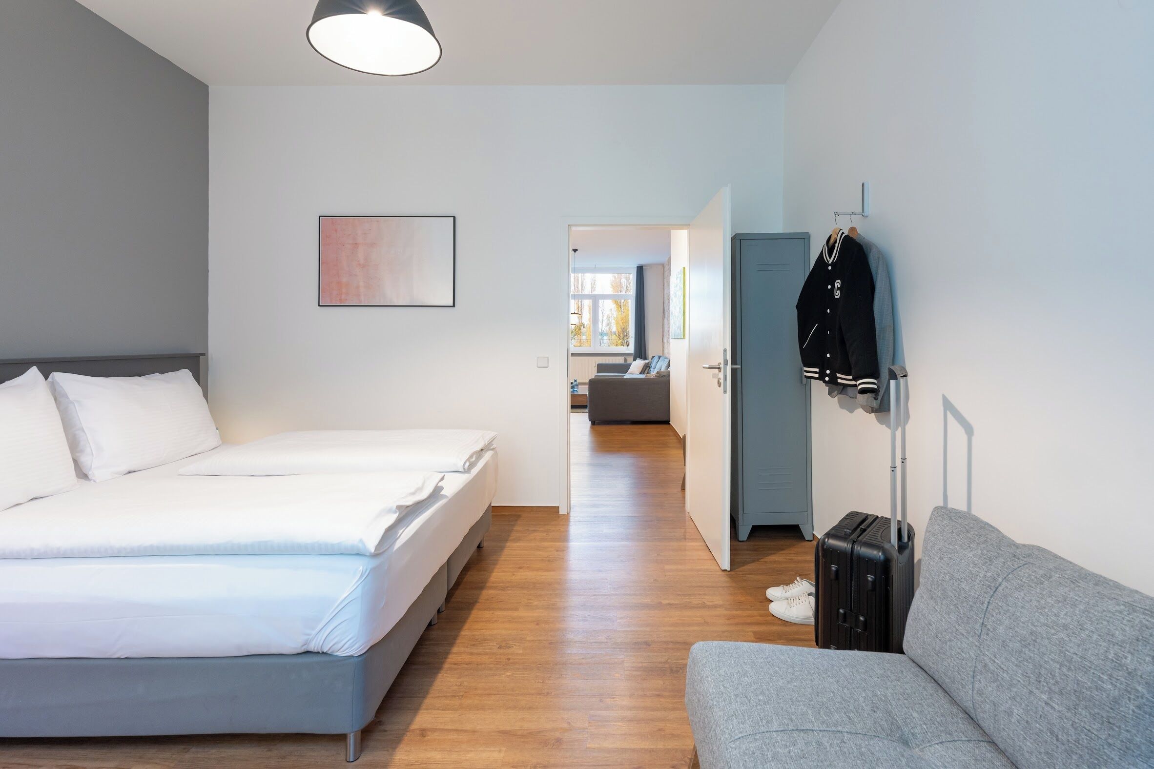 Quarto de hotel moderno com uma cama grande, um sofá cinzento, uma mala e um porta-casacos.