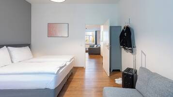 Quarto de hotel moderno com uma cama grande, um sofá cinzento, uma mala e um porta-casacos.