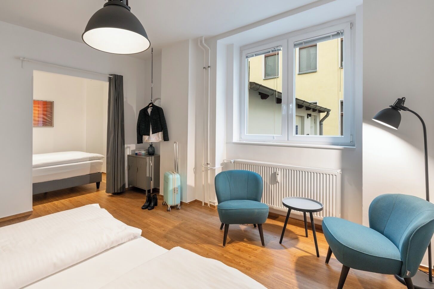 A modern hotel room with a bed, two chairs, a small table, a suitcase, a coat rack, and a window with a view of a building.