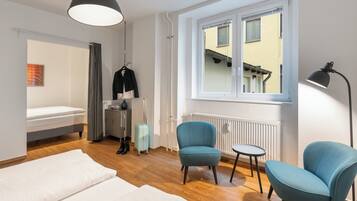 A modern hotel room with a bed, two chairs, a small table, a suitcase, a coat rack, and a window with a view of a building.