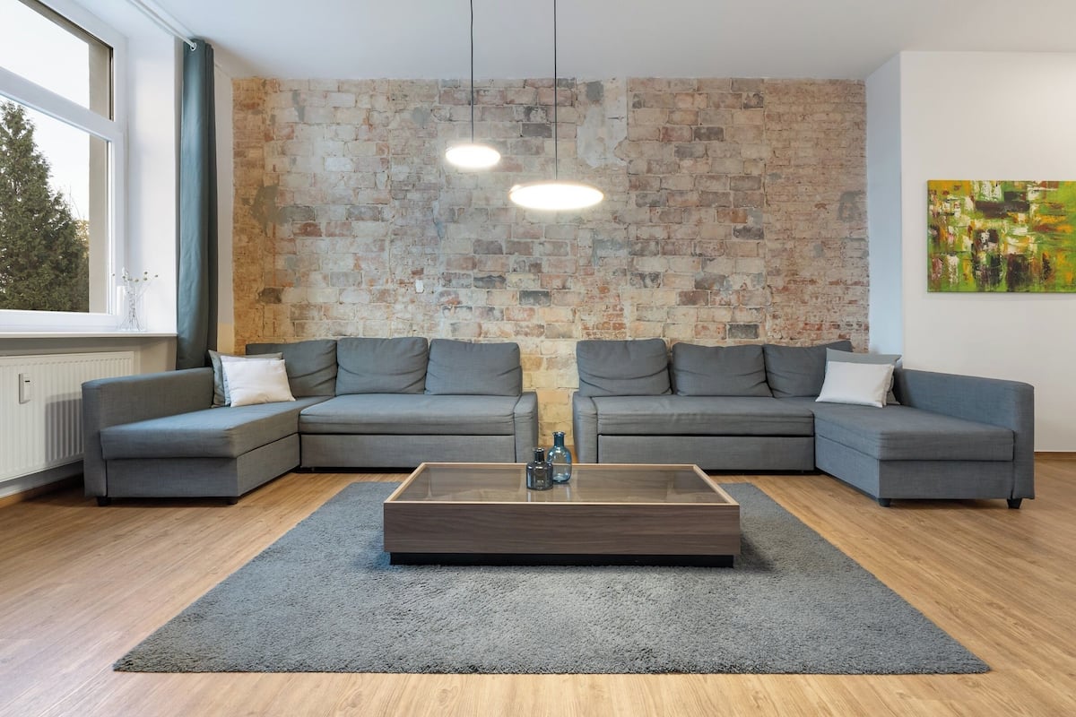 A modern living room with a sectional sofa, a wooden coffee table, and a brick wall.