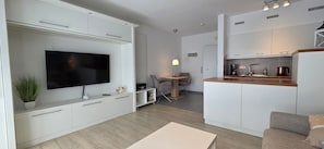 A modern living room with a flat-screen TV, a dining area with a table and chairs, and a kitchen with white cabinets and a wooden countertop.