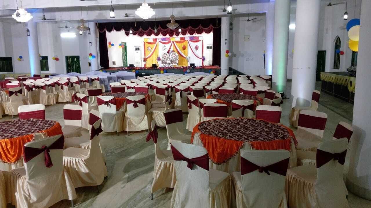 Un salón amplio con filas de mesas y sillas, un escenario con un telón de fondo y columnas.