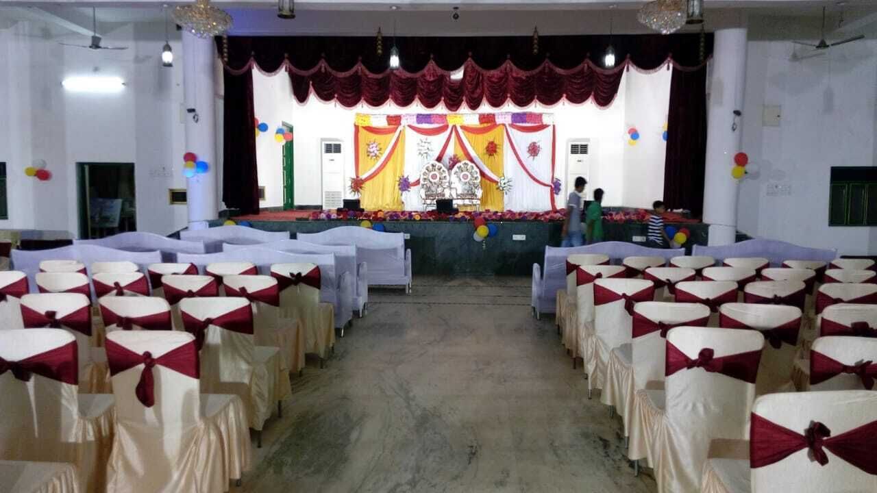 Un salón con filas de mesas y sillas, un escenario con fondo decorativo y una araña.