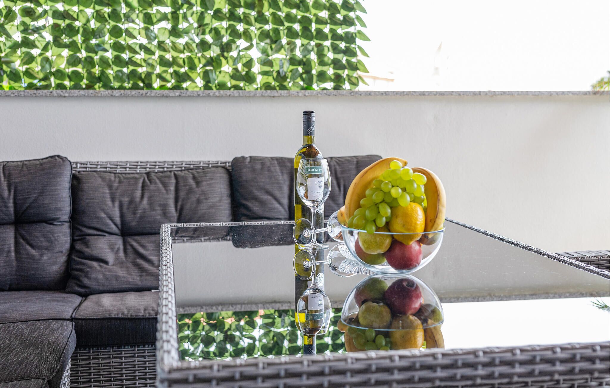 A glass table with a bottle of wine, a wine glass, and a fruit bowl containing grapes and apples.