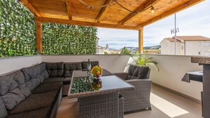 Ein überdachter Außenbereich mit einer Couch und Korbstühlen, einem Glastisch mit Früchten und Blick auf Gebäude im Hintergrund.