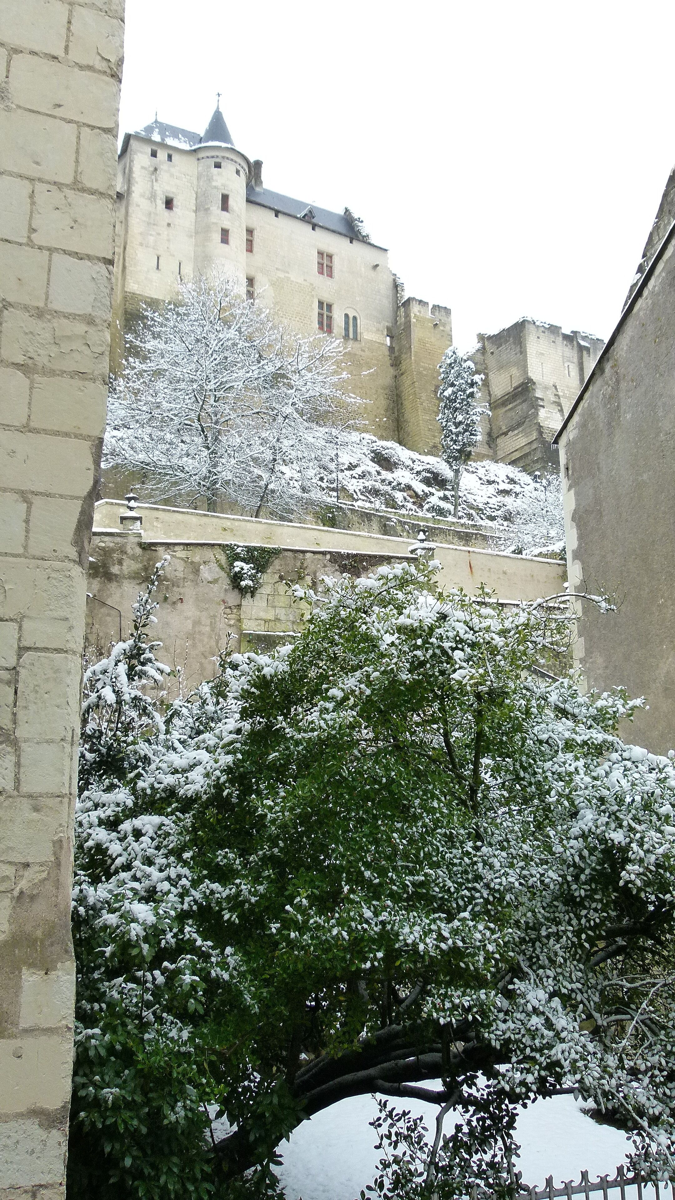 Une cour enneigée avec un pont de pierre, un grand bâtiment doté d’une tour et un mur percé d’une fenêtre.
