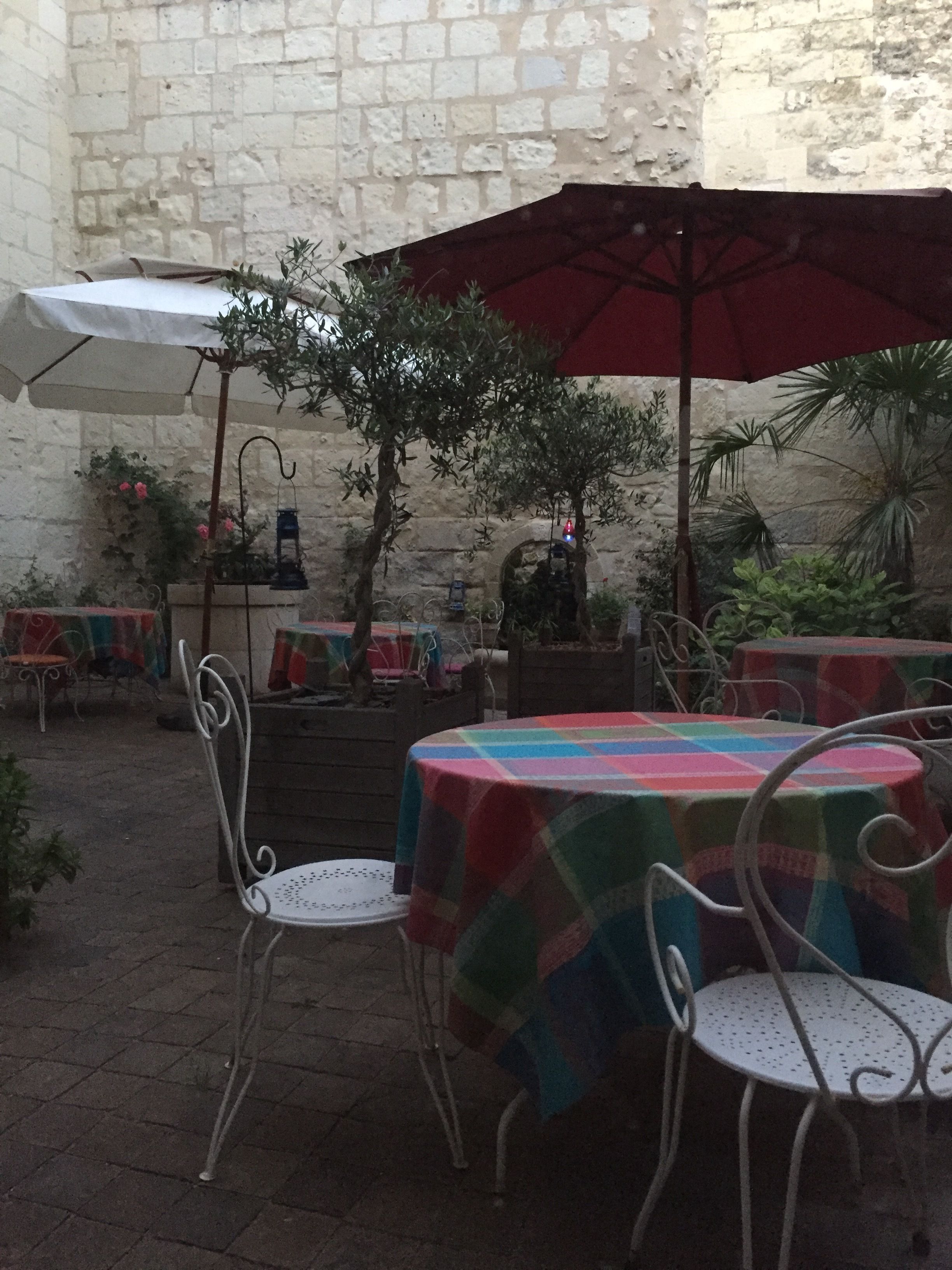 Un patio al aire libre con paredes de piedra, mesas con manteles de colores, sillas de metal y sombrillas.