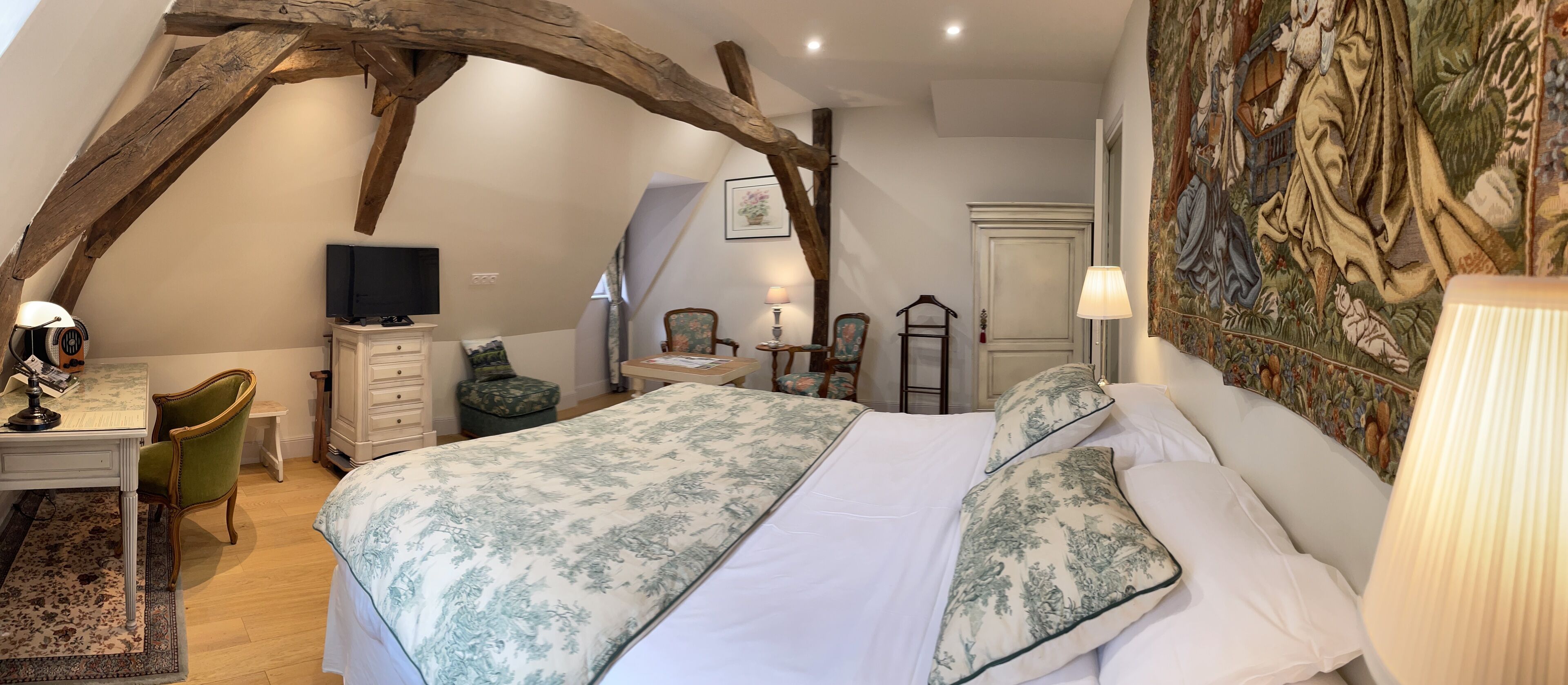 A bedroom with a large bed, a desk with a chair, a television, and a tapestry on the wall.