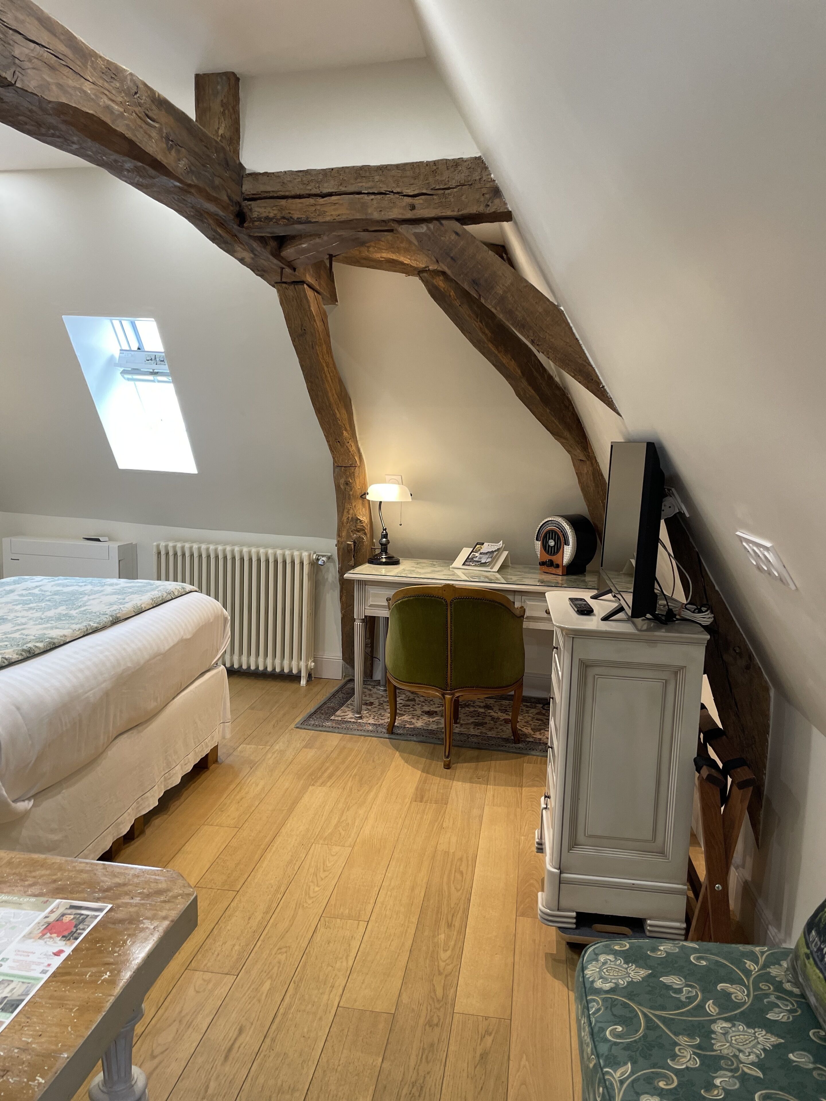 A bedroom with a loft-style ceiling, a bed, a desk with a chair, a radiator, and a window.