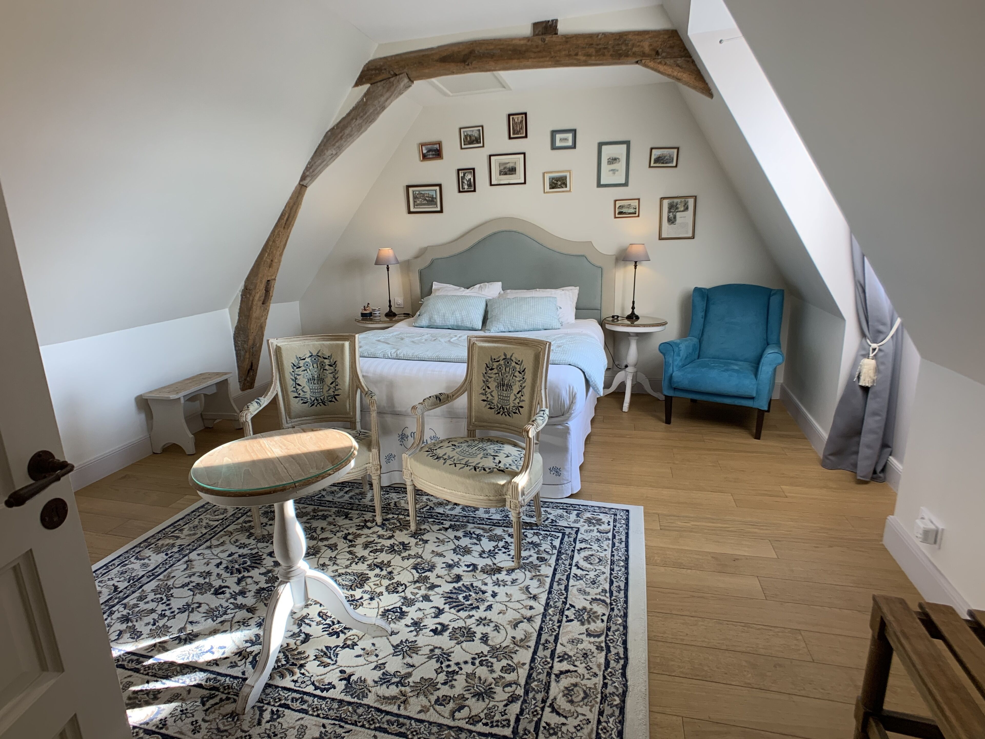 A bedroom with a bed, two chairs, a small table, a blue armchair, a patterned rug, and a wooden ceiling beam.