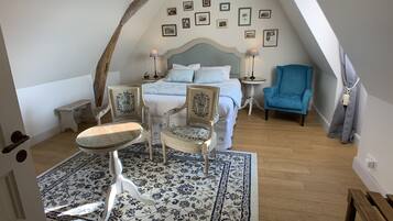 A bedroom with a bed, two chairs, a small table, a blue armchair, a patterned rug, and a wooden ceiling beam.