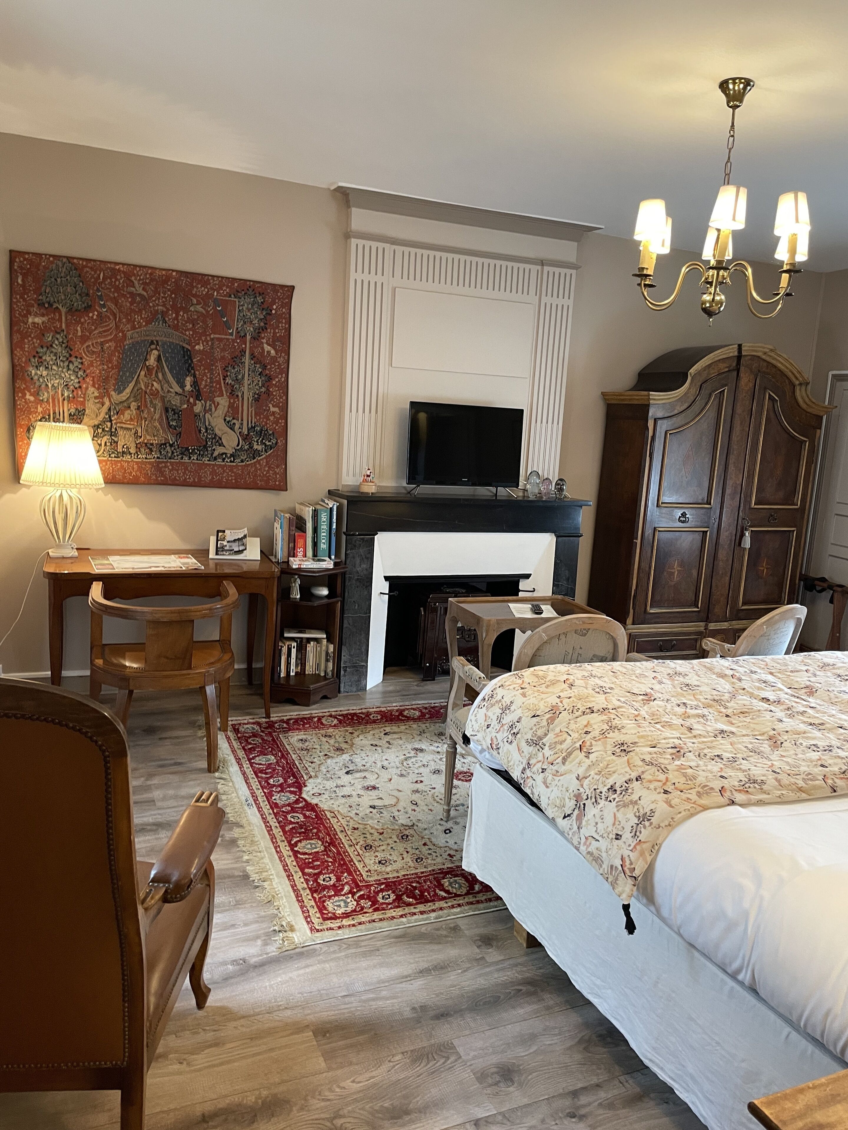 A hotel room with a bed, a desk, a television, a fireplace, a wardrobe, and a large tapestry on the wall.