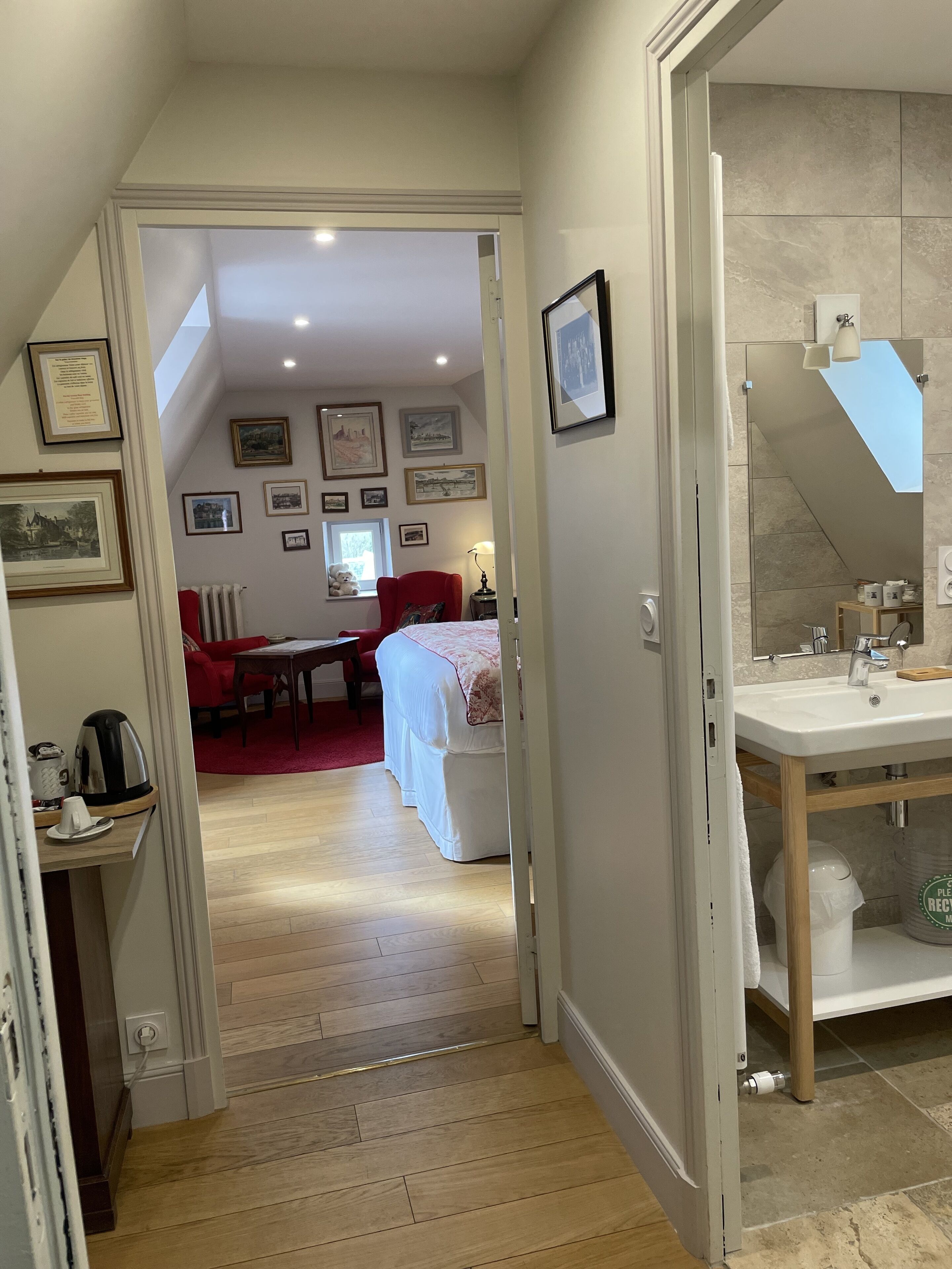 A hallway leading to a bedroom with a red chair, a bed with white bedding, and a bathroom with a sink and mirror.