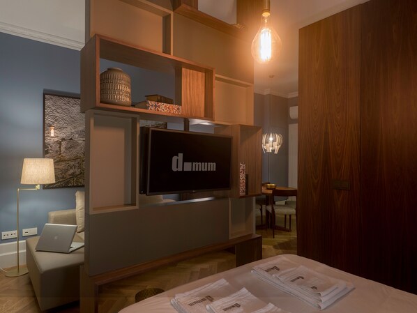 A modern hotel room with a flat-screen TV mounted on a wooden wall, a dining area with a chandelier, and a bed with neatly folded towels. - Domum 2 (Porto)