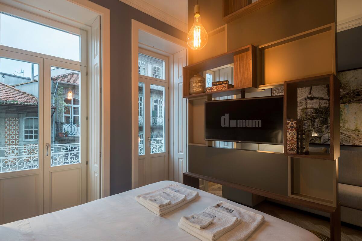 Quarto de hotel com uma cama, duas janelas, uma televisão e uma estante decorativa.