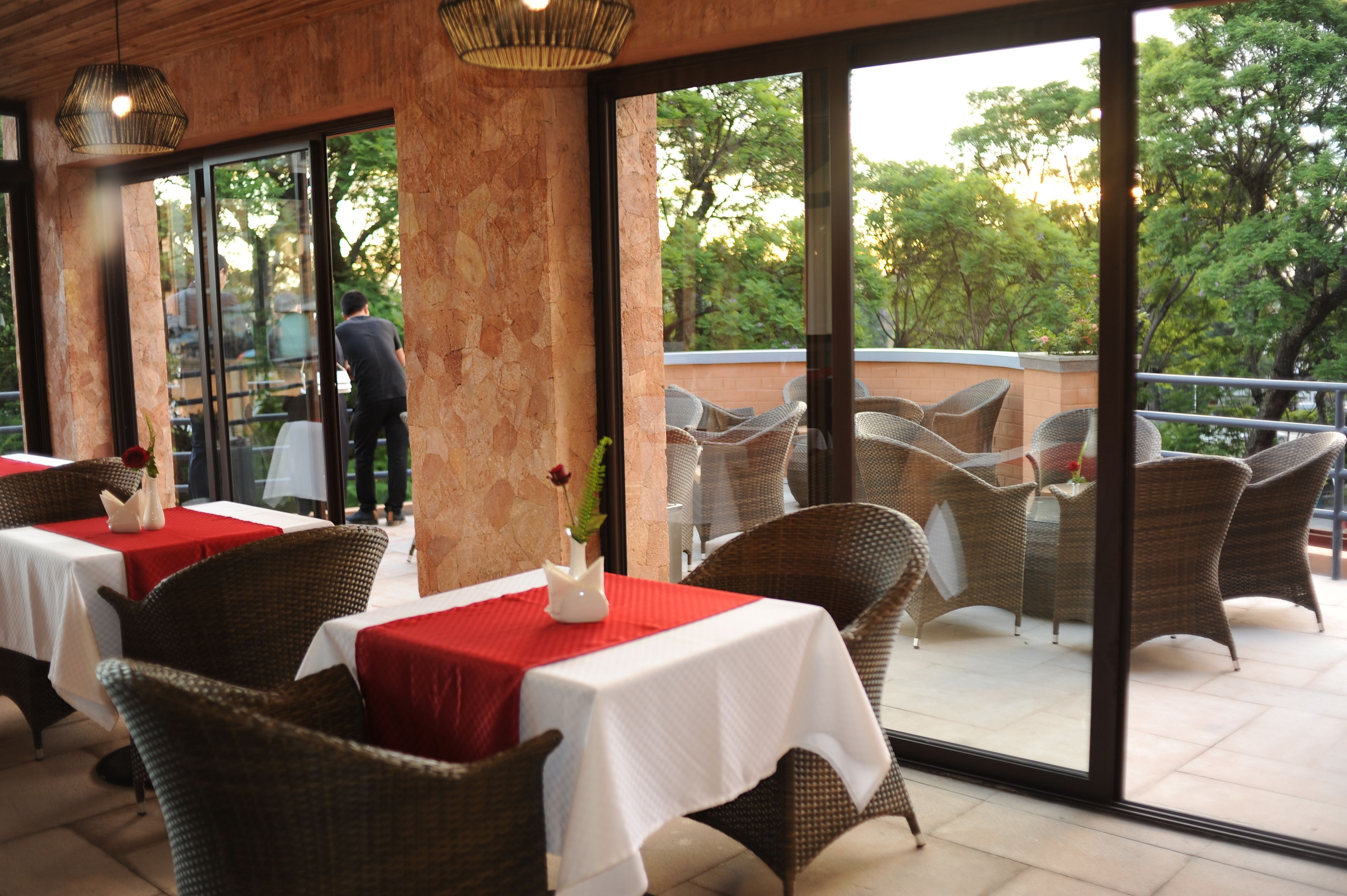 Un área de comedor al aire libre con sillas y mesas de mimbre, puertas de vidrio grandes que dan a un balcón con vistas a árboles.