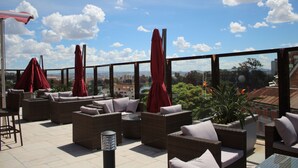A rooftop terrace with wicker furniture, red umbrellas, and a city view. - Havana Resort (Antananarivo)