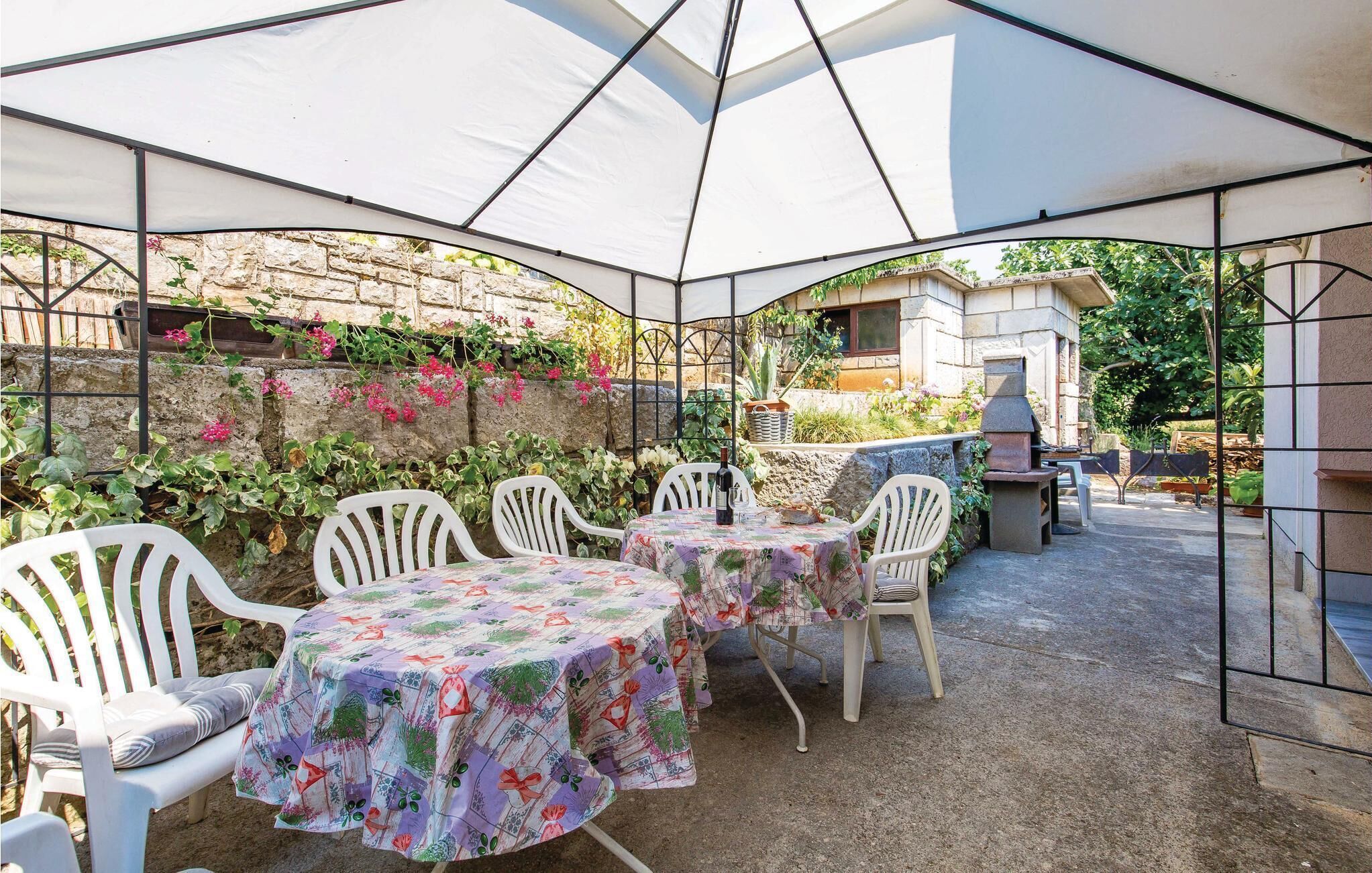 Une terrasse extérieure avec des chaises et des tables en plastique blanc recouvertes de nappes à motifs floraux, un mur de pierre orné de plantes grimpantes et un four en pierre.
