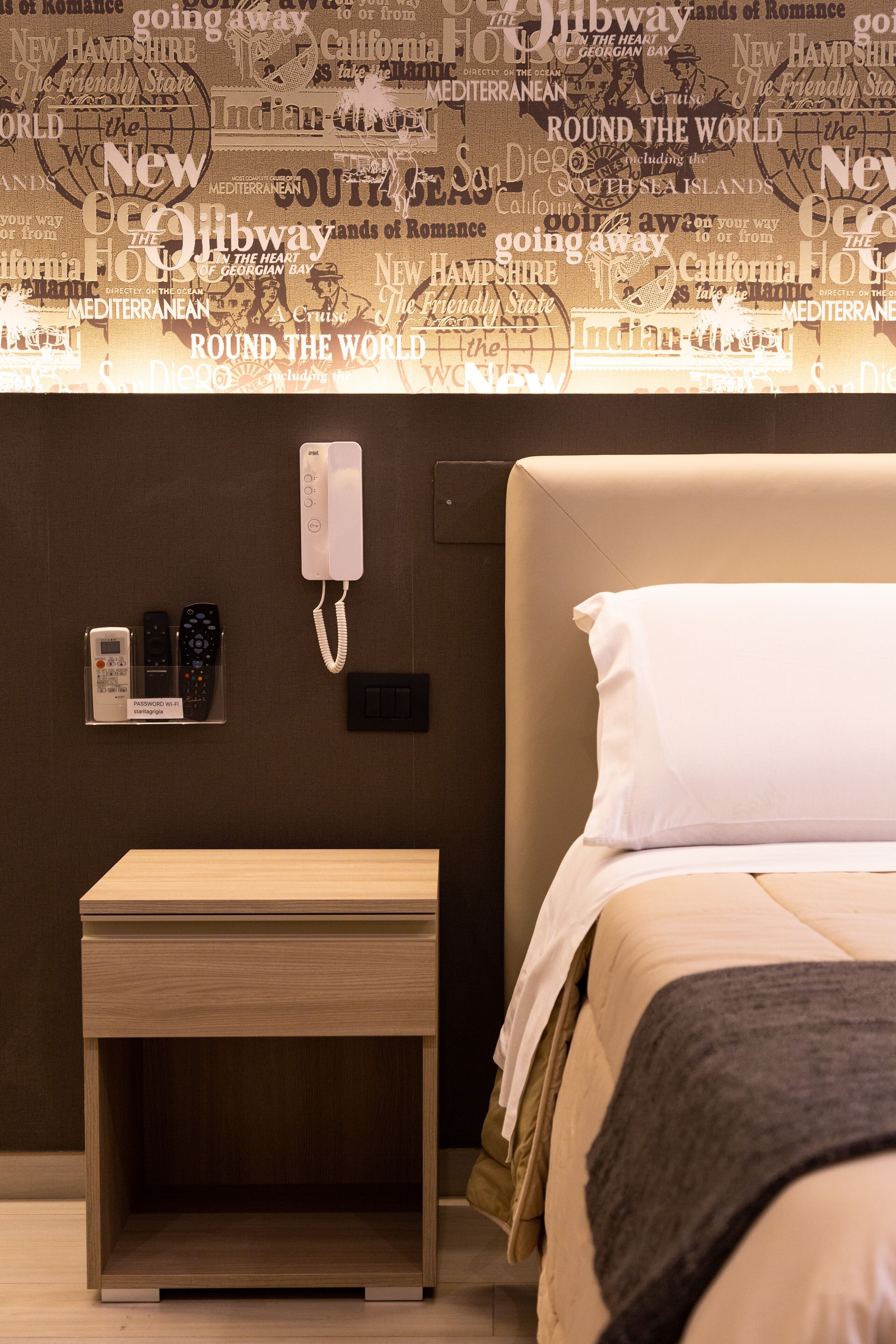 A hotel room with a bed, a bedside table, a phone, and a wall with travel-related text.