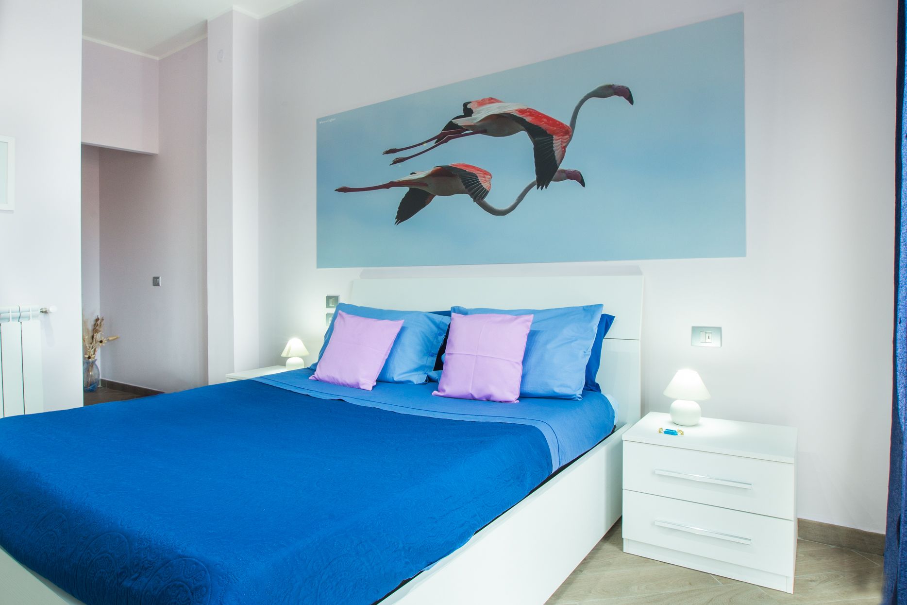 A bedroom with a large bed, a nightstand, a wall-mounted light, and a painting of flamingos.