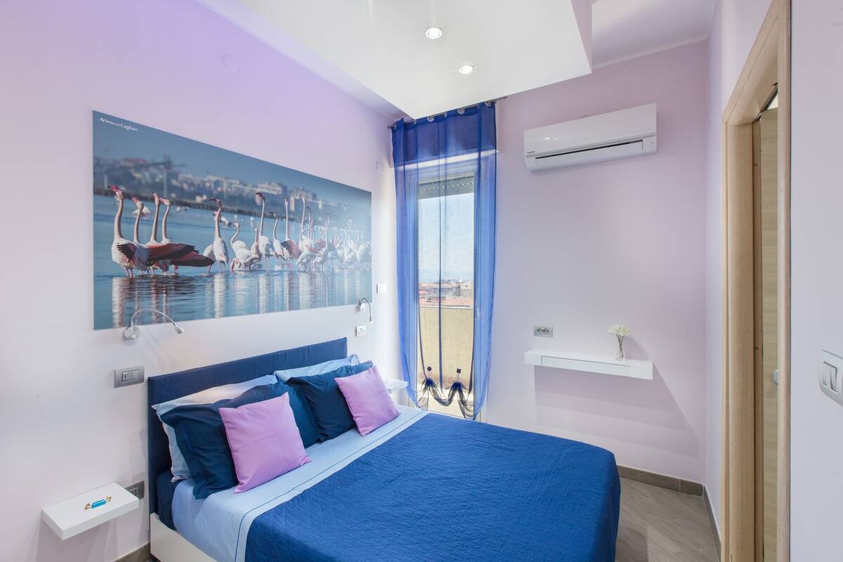 A bedroom with a bed, a painting of swans, a window with blue curtains, and an air conditioning unit.