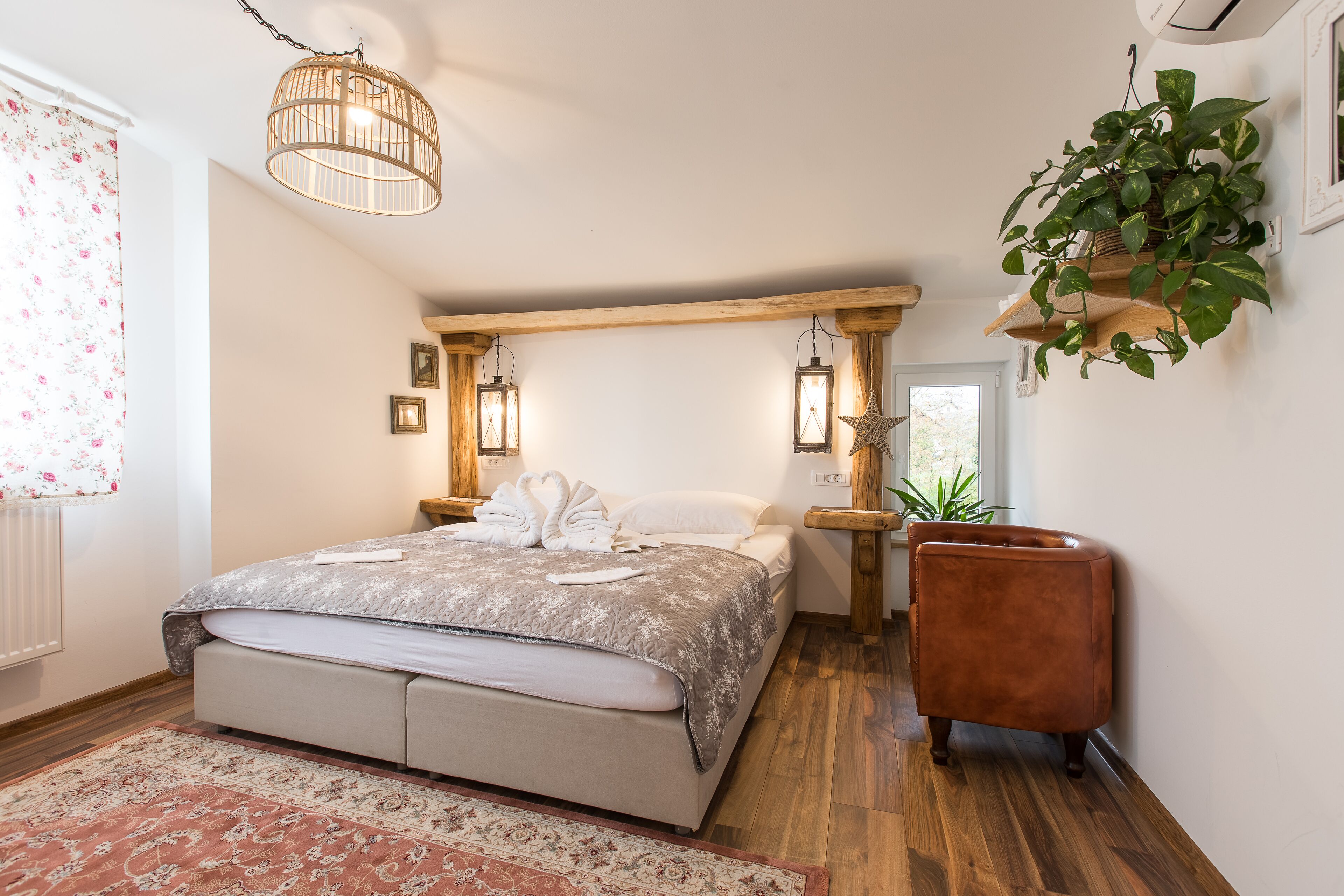 A bedroom with a large bed, wooden beams, a hanging plant, and a small brown chair.