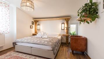 A bedroom with a large bed, wooden beams, a hanging plant, and a small brown chair.