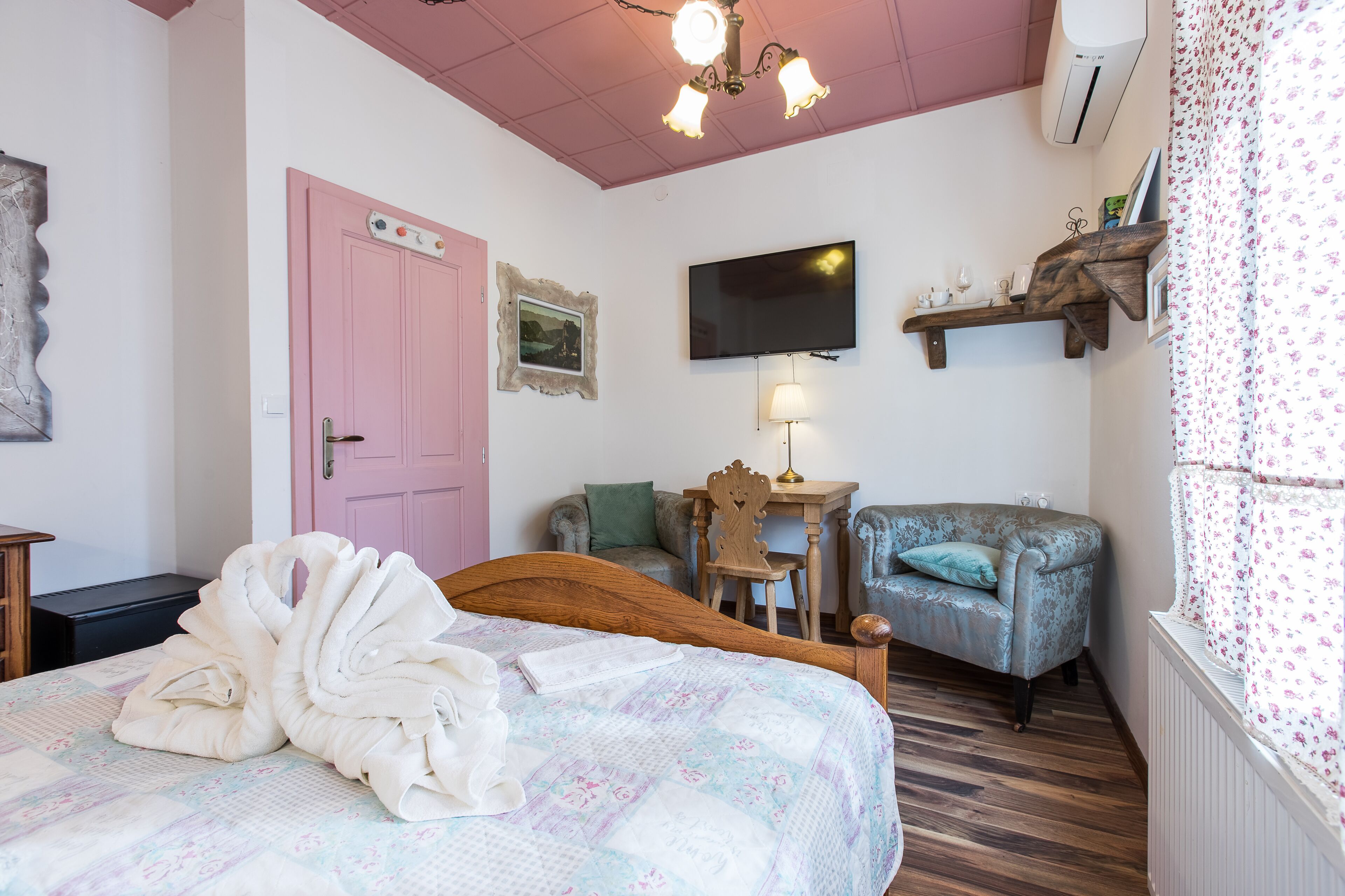 A bedroom with a bed, a pink door, a television, a wooden chair, a table, and a window with floral curtains.