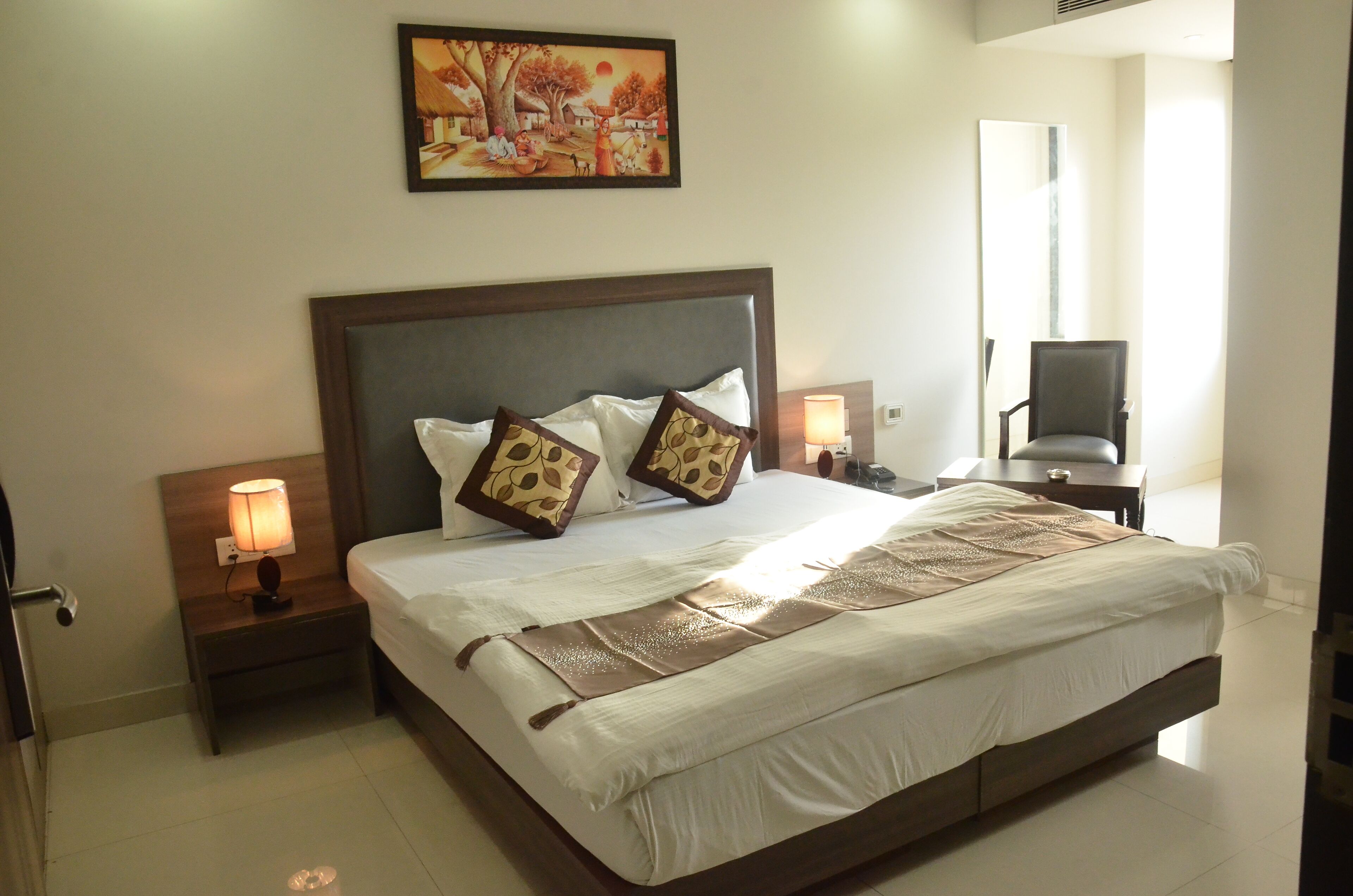 A neatly made bed with a headboard, two bedside tables with lamps, a chair, and a painting on the wall.