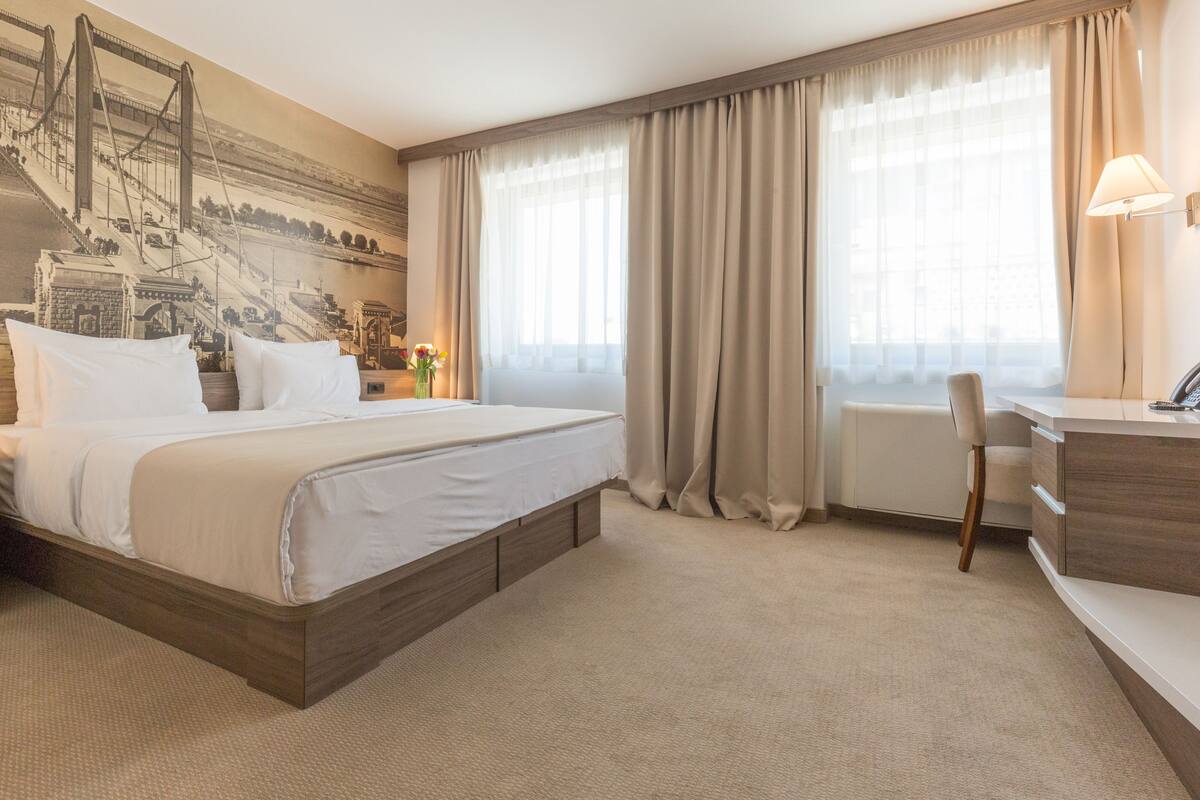 A hotel room with a large bed, a desk, a chair, and a window with curtains.