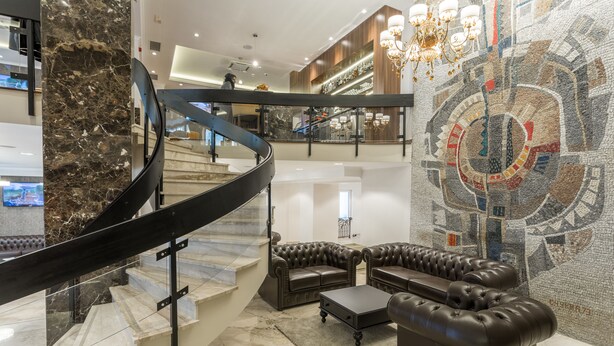 A modern lobby with a marble staircase, leather seating, and a mosaic wall. - Hotel Royal Inn (Belgrade)