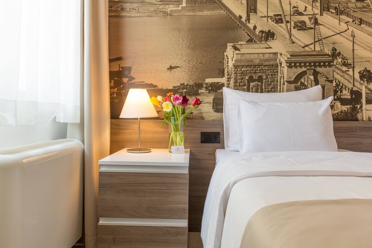 A hotel room with a bed, a nightstand with a lamp, a vase of flowers, and a mural on the wall.