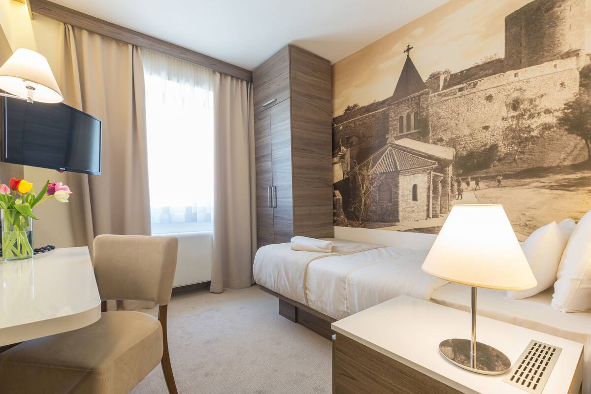 A hotel room with a bed, a desk with a chair, a TV, and a painting of a historical building.