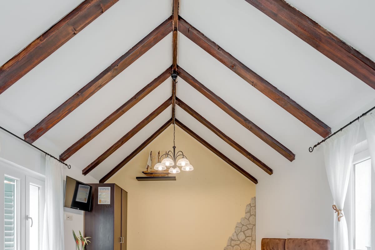 A bedroom with a sloped ceiling, wooden beams, a bed with white bedding, a small bedside table, and a TV mounted on the wall.